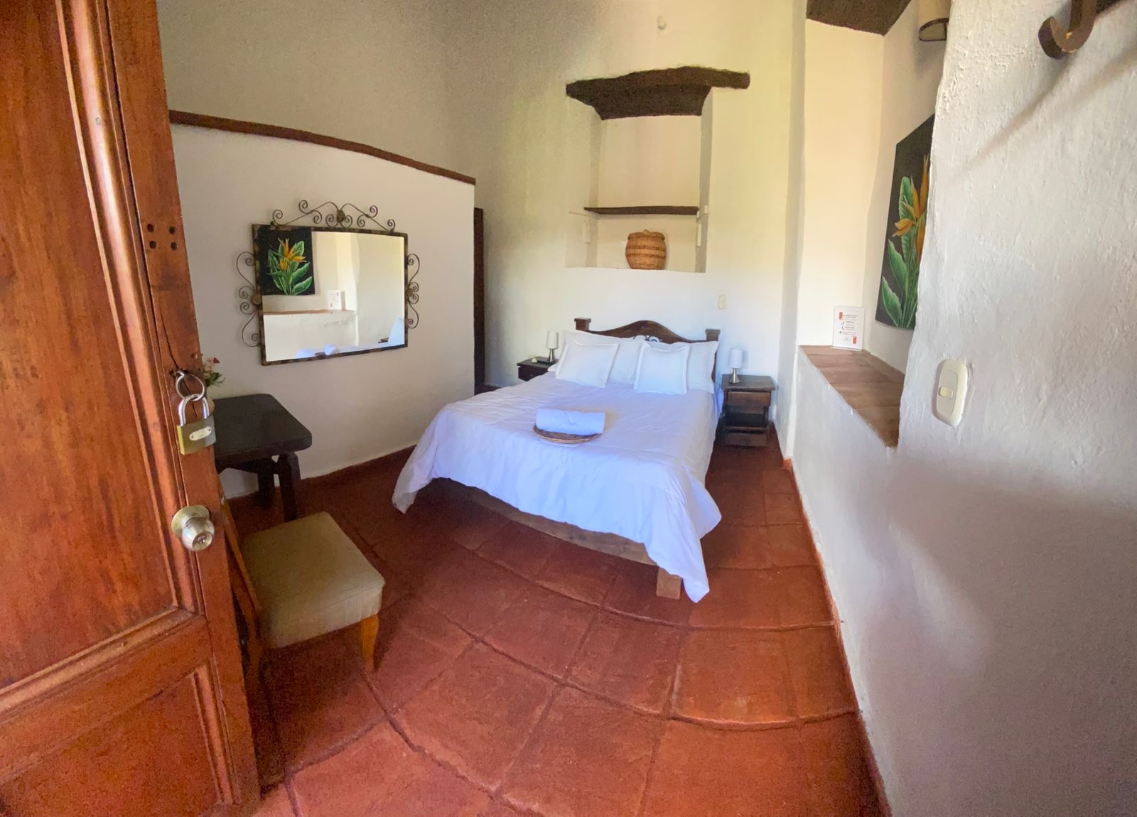 Habitación sencilla con cama doble, escritorio, espejo de forja y piso de baldosa de barro.